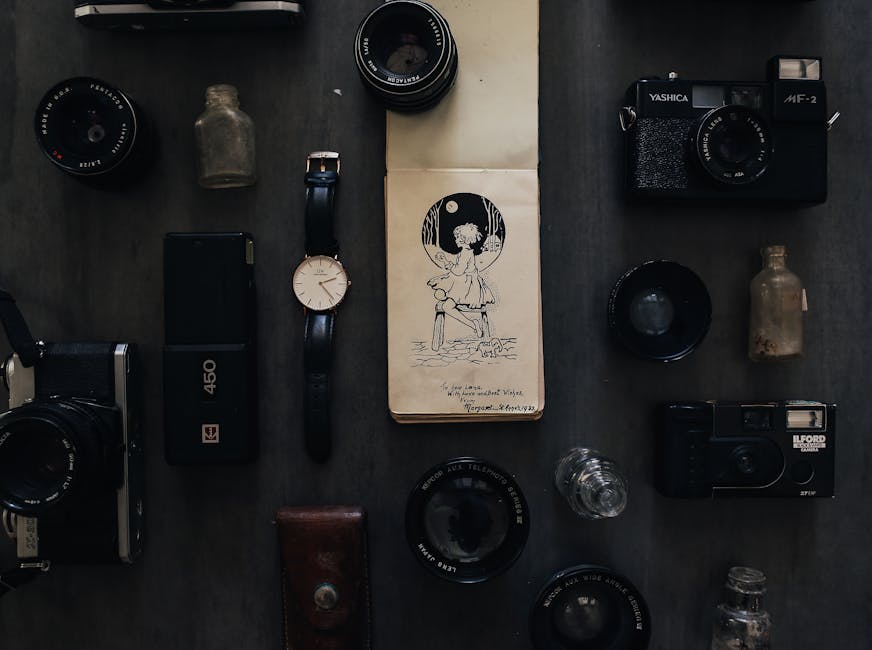 An overhead view of various personal items and electronics arranged on a dark surface, including a black and silver film camera, a black digital camera with a leather strap, a digital voice recorder, a wristwatch with a black strap and white face, a small glass bottle, a smartphone, a blue lens, and multiple camera lenses of different sizes and focal lengths. There are also small glass bottles, a leather pouch, and a paper notebook with a black and white illustration of a girl sitting on a bench under a tree, with handwritten text at the bottom. The items appear to be packed for a home relocation, with some objects positioned near the edge of the surface as if ready for loading into a vehicle. The setting suggests a preparation or packing process related to furniture transport and moving logistics, as handled by Ilford Movers, with the focus on careful packing and organization for a house removal.