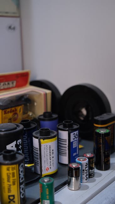 Close-up image of a collection of various AA and AAA batteries, some with clear labels indicating voltage and type, arranged on a smooth surface likely indoors. In the background, blurred objects are visible, including black speakers, a cardboard box, and packaging materials. The scene appears to be part of a home environment, possibly a storage or packing area used during a house removal, with the battery collection ready for packing or transport. The lighting is soft and evenly distributed, highlighting the details of the batteries and surrounding items. This image illustrates the careful handling and organization of small household items during a furniture transport or packing process, consistent with services offered by Ilford Movers for comprehensive home relocation, ensuring all items, including electronics and batteries, are properly packed and protected for transport.
