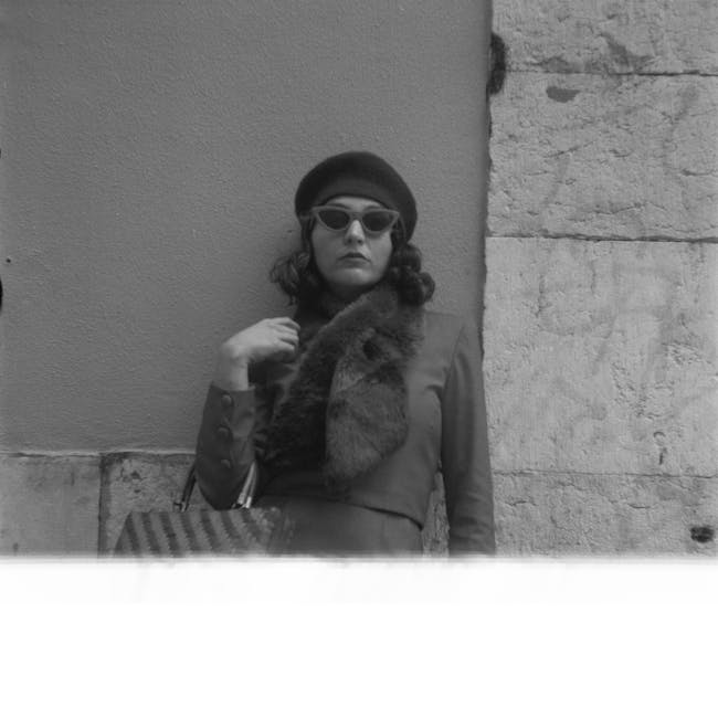 A woman in transit standing beside a stone and plaster wall, possibly outside a building designated for house removals or moving services. She is wearing dark sunglasses, a dark beanie, and a coat with a fur collar, holding a small handbag with a striped pattern. The setting suggests a professional packing and moving context, with her posture indicating readiness for furniture transport or home relocation. The photograph is in black and white, emphasizing the environment and attire, and subtly aligns with content about removals, packing, and logistics services offered by Ilford Movers, relevant to the IG1 area and the detailed parking and rates information on the webpage for Ilford Town Centre.