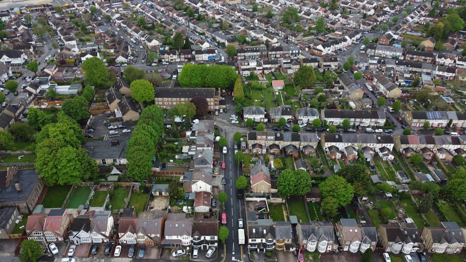 An aerial view of a residential neighbourhood showing densely packed houses with varied roof styles, surrounded by green trees and small gardens. The roads are narrow, lined with parked cars, and some houses have small front yards with hedges and fences. The scene captures a typical suburban area during daylight hours, with natural lighting highlighting the rooftops and foliage. In the foreground, a section of street is visible with parked vehicles and the end of a driveway leading to a house, indicating a home relocation or furniture transport process supported by Ilford Movers. The image emphasizes the compactness of the housing estate, suitable for illustrating house removals, packing, and loading activities within a typical property environment for Ilford removals services.