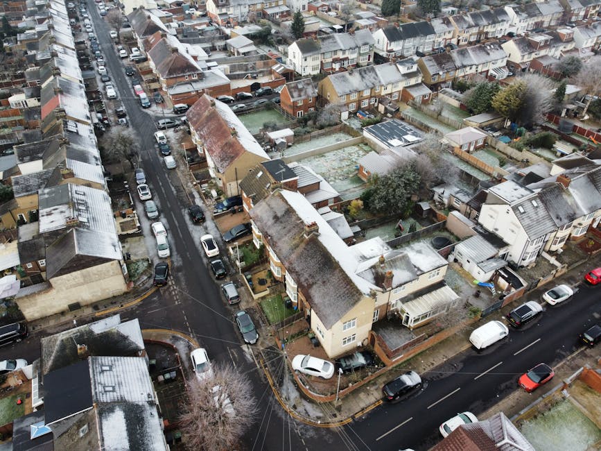 An aerial view of a residential neighbourhood showing a row of semi-detached houses with pitched roofs, some covered in snow, and small back gardens. The street is lined with parked cars on both sides, and a narrow road intersects the scene, with vehicles parked along its curb. Inside one of the back gardens, a crew from Ilford Movers is engaged in a home relocation process, using a trolley and blankets to carefully lift and load a piece of furniture, possibly a sofa or large box, towards a nearby moving van parked on the street. The van's sliding door is open, revealing packing materials like cardboard boxes and plastic wrap inside, indicating ongoing packing and furniture transport preparations. The surroundings feature trees, small sheds, and fencing, with daylight illuminating the scene, consistent with a typical house removal operation in Cranbrook Road or nearby estates in Ilford.