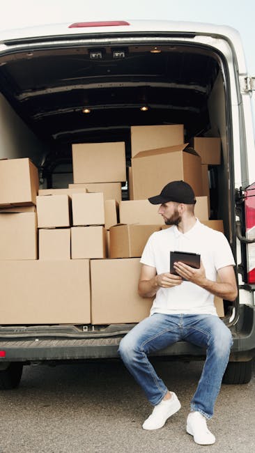 A man standing outside a residential property next to a white moving van with its rear doors open, engaged in the packing and loading process for home relocation. The man is holding a cardboard box, w