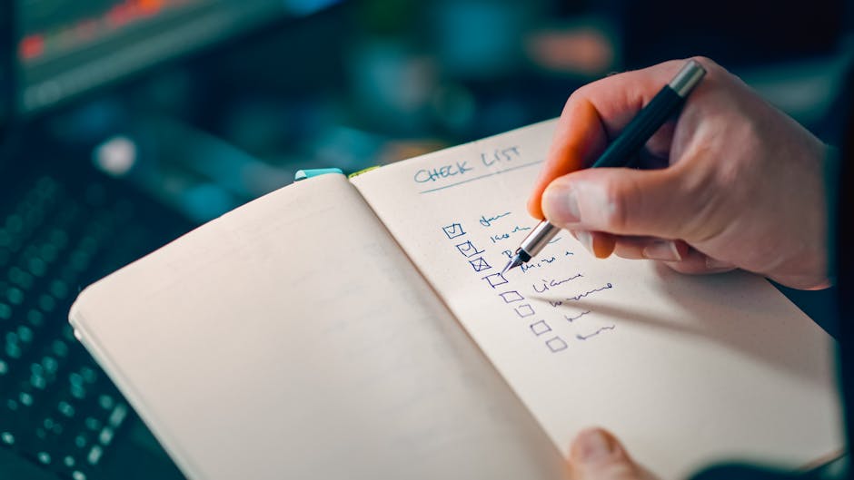 A close-up of a person's hand holding a silver pen, writing a checklist titled 'CHECK LIST' in a small, spiral-bound notebook with cream-colored pages. The checklist contains several itemized tasks, each marked with a small square checkbox, some checked off and others blank. The background includes a partially visible computer keyboard with illuminated keys, suggesting an office environment, where the individual is likely preparing or reviewing tasks related to a house or office relocation. The scene emphasizes planning and organizing efforts typical during a professional removal or moving process, such as packing, transport, and logistics management, with the notation indicating detailed pre-move preparations carried out by Ilford Movers in the context of office removals.