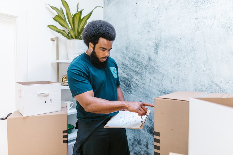 A professional mover from Ilford Movers, dressed in a navy blue t-shirt, is inspecting a packing checklist on a clipboard while standing inside a property during a house relocation. The room features a textured light blue wall and a white shelf holding a large potted plant with long, green leaves, a white mug, and some small items. Several cardboard boxes of various sizes, some sealed with packing tape and others open, are stacked around the mover, indicating packing and moving preparations. The boxes are made of brown corrugated cardboard, with a white box on top of one of the stacks. The mover appears focused, possibly coordinating the loading process or verifying items for transport. Natural light illuminates the scene, suggesting daytime. This image exemplifies the detailed planning involved in furniture transport and packing during relocation services, with Ilford Movers facilitating a smooth household moving process.