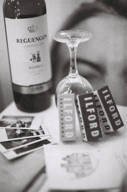 A close-up black and white photograph showing a wine bottle with a label reading 'Requengo,' placed upright behind an upside-down wine glass that is centered in the image. In the foreground, a small stack of printed photographs is spread out on a flat surface. To the right of the photograph, there are two overlapping rectangular labels with bold text reading 'ILFORD' and 'LIFE FORD,' which are partially curved and positioned adjacent to the wine glass. The background appears blurred, suggesting an indoor setting. The image does not depict any active moving or packing process but visually highlights items that could be associated with domestic or social activities.