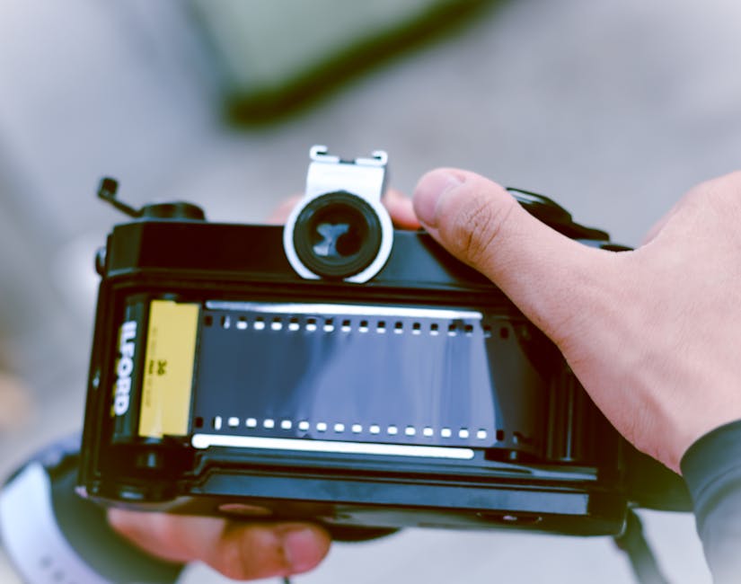 A person holding a vintage medium format film camera, viewed from above, with the camera body showing a yellow film insert on the left side and an empty film compartment with sprocket holes visible. T