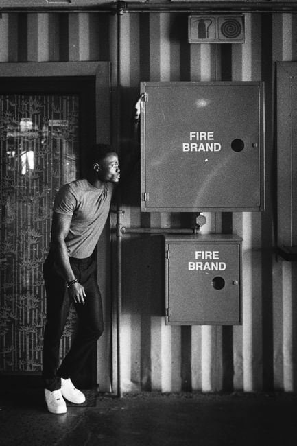 A young man dressed in a casual t-shirt, trousers, and white sneakers stands indoors next to a large, grey fire safety cabinet mounted on a wall with vertical striped paneling. The cabinet is labeled 'FIRE BRAND' and has a smaller compartment below it. The man is reaching up with his right hand towards the cabinet door, appearing to check or operate it, while his left hand rests at his side. To the left of the man, there is a decorative glass door with a patterned design, allowing some light to pass through. The environment is dimly lit, emphasizing the utilitarian nature of the setting. This scene depicts a typical safety or maintenance check, relevant to building safety procedures involved in house removals or property preparation in locations such as Seven Kings, Ilford. Ilford Movers, a professional removals company, operates within this context of moving and packing logistics, ensuring safety compliance during home relocations.