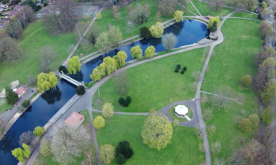 An aerial view of Valentines Park and the surrounding Redbridge area in Ilford, showing lush green open spaces, walking paths, and scattered trees. In the foreground, the park features grassy fields w