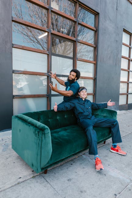 Two men involved in a home relocation are positioned outside a building with large, grid-patterned windows and a grey facade. One man, standing behind a green velvet sofa, holds a cardboard box and appears to be lifting or moving it toward the building entrance. The second man, seated on the sofa, is smiling, extending his arms outward, and dressed in casual dark clothing with bright red sports shoes. The sofa is situated on a concrete sidewalk, with the men demonstrating an active moving process as part of furniture transport during packing and moving activities. The building's windows reflect trees and the sky, indicating an outdoor setting associated with relocating belongings to or from the property. The scene captures a professional yet relaxed moment in the logistical process of house removals, aligning with the services offered by Ilford Movers for residential moves in the Ilford area, including parkside relocations near Valentines Park.