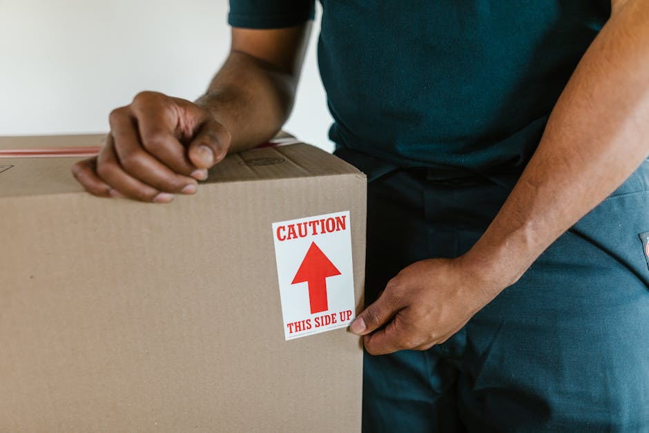 A person in a dark green shirt and dark trousers is shown during a home relocation process, gripping the edges of a large cardboard moving box that has a red and white caution label marked with an upward arrow and the instruction 'This Side Up'. The individual is in the act of either preparing to load or unload the box, which is situated indoors on a floor with a visible strip of carpet or flooring alongside the wall. Surrounding the box are other packing materials, including plastic wrap or packing paper, indicating careful packing for furniture transport. The lighting suggests an indoor environment, and the focus is on the careful handling of the box within a moving context, emphasizing the importance of proper packing and logistics in house removals, as handled by companies like Ilford Movers. This visual relates directly to the process of packing and loading during a professional house move, supporting the theme of relocations and transportation services in the Ilford area, in accordance with the 'Valentines Park movers insider tips for Parks​ide moves' page.
